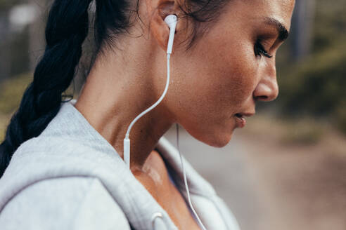 Close up of runner with sweat standing outdoors after her workout. Tired fitness female with earphones resting after exercising outdoors. - JLPSF14912