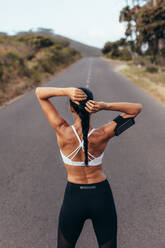 Rückansicht einer fitten jungen Frau, die sich die Haare zusammenbindet und sich auf ein Lauftraining vorbereitet. Eine Sportlerin vor einem Lauf auf einer leeren Straße. - JLPSF14926