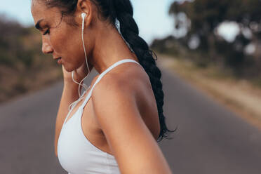Seitenansicht einer Fitness-Frau, die auf einer leeren Straße steht. Frau in Fitnesskleidung, die auf einer Straße steht und mit Kopfhörern Musik hört. - JLPSF14930