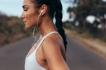 Seitenansicht einer Läuferin, die morgens auf einer Straße steht. Eine Frau in Fitnesskleidung steht auf einer Straße und hört mit Kopfhörern Musik. - JLPSF14931