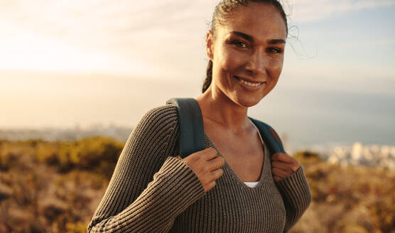 Happy woman carrying a bag walking through field. Young female on country hike, looking at camera and smiling. - JLPSF14957