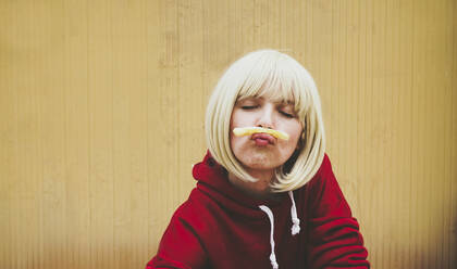 Frau mit geschlossenen Augen balanciert Pommes frites auf den Lippen vor einer braunen Wand - SVCF00199