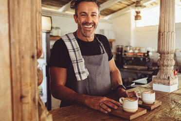 Männlicher Barista, der den Kunden Kaffee serviert. Mittlerer erwachsener Mann mit Schürze, der mit zwei Tassen Kaffee auf dem Tresen in die Kamera schaut. - JLPSF14981