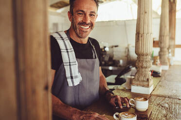 Mittlerer erwachsener Mann in Schürze schaut in die Kamera und lächelt, während er hinter der Theke steht. Lächelnder männlicher Barista in einem Café. - JLPSF14982