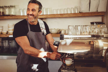 Männlicher Barista, der in einem Café einen Espresso zubereitet, wegschaut und lächelt. Mann in Schürze, der in einem Café mit einer Kaffeemaschine Kaffee zubereitet. - JLPSF14984