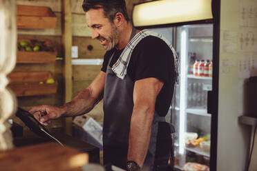 Male barista at counter using cashbox computer in cafe store checking client's order. Waiter in apron working in coffee shop. - JLPSF14986