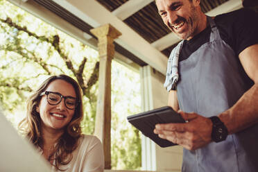 Young woman in a cafe ordering from menu while waiter taking order using a digital tablet. Smiling waiter in apron standing at table and adding information into tablet while taking order from guest in cafe. - JLPSF14991