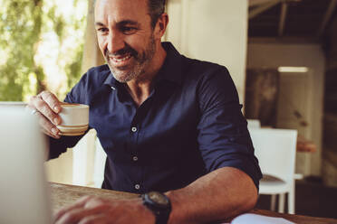 Lächelnder Geschäftsmann, der einen Laptop benutzt und Kaffee trinkt, während er am Tisch in einem Café sitzt. Ein Mann trinkt eine Tasse Kaffee und schaut auf seinen Laptop. - JLPSF14996