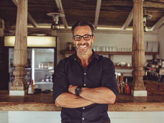 Portrait of a successful coffee shop owner standing at the cafe counter. Handsome man standing with his arms crossed at his own restaurant. - JLPSF15000