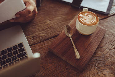 Top view of cup of hot cappuccino art coffee on wooden table with a man reading document. Cup of coffee with milk foam art pattern on cafe table with laptop on side. - JLPSF15004