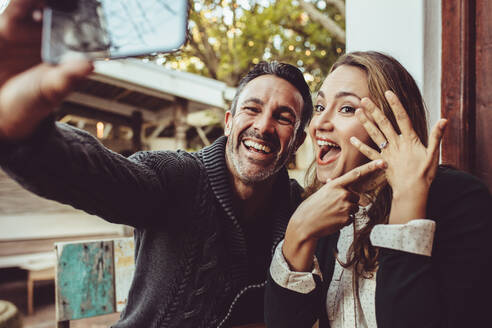 Zärtliches Paar, das seine Verlobung mit Selfies bekannt gibt, während es im Café sitzt. Glückliches Paar, das ein Selfie macht und seinen Ehering im Café zeigt. - JLPSF15016