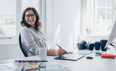 Professionelle Fotografin sitzt an ihrem Bürotisch, schaut weg und lächelt. Frau im Büro mit digitalem Grafiktablett und Zeichenstift. - JLPSF15023