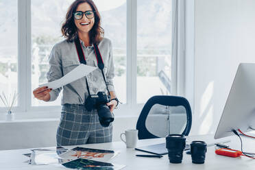 Young woman photographer standing in studio. Smiling female checking printed images at her office desk. - JLPSF15024