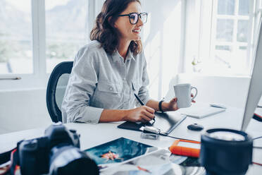 Junge Frau sitzt am Schreibtisch und bearbeitet Bilder am Computer. Fotografin retuschiert Fotos im Büro mit Grafiktablett und digitalem Stift. - JLPSF15036