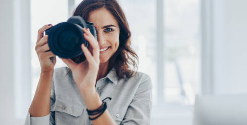 Glückliche junge Frau, die mit ihrer DSLR-Kamera drinnen Fotos macht. Professionelle Fotografin, die Fotos in ihrem Studio macht. - JLPSF15044