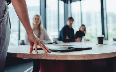 Weibliche Führungskraft, die am Tisch steht und sich an ihr Team wendet, das im Sitzungssaal sitzt. Fokus auf die Hand der Geschäftsfrau am Tisch, während sie mit Kollegen im Sitzungssaal spricht. - JLPSF15135