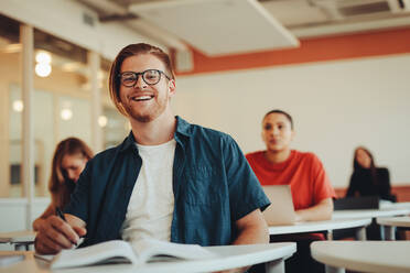 Porträt eines jungen männlichen Schülers im Klassenzimmer der High School. Junger Mann im Klassenzimmer eines Kollegen, der in die Kamera schaut und lächelt. - JLPSF15142