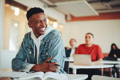 Männlicher Student, der in einem Klassenzimmer der Universität sitzt, wegschaut und lächelt. Mann sitzt in einer Vorlesung in einem Klassenzimmer der High School. - JLPSF15145