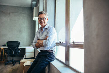 Smiling businessman looking at camera as he stands by a window sill with arms crossed. Mid adult businessman at his office. - JLPSF15176