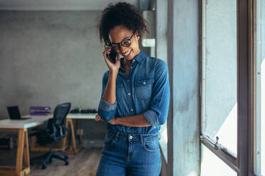 Positive young woman in casuals talking on mobile phone. Smiling female entrepreneur standing by a window in office and talking over cell phone. - JLPSF15180