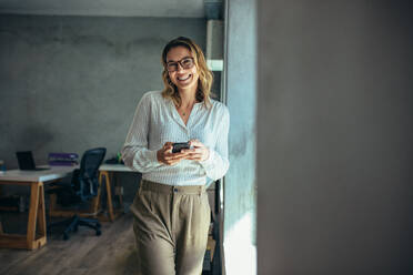 Cheerful woman standing in office with her phone. Businesswoman with cell phone in hand looking at camera and smiling. - JLPSF15187
