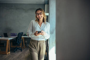 Fröhliche Frau steht mit ihrem Telefon im Büro. Geschäftsfrau mit Handy in der Hand schaut in die Kamera und lächelt. - JLPSF15187