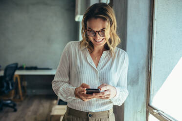 Geschäftsfrau, die ihr Smartphone im Büro benutzt. Unternehmerin, die auf ihr Mobiltelefon schaut und lächelt. Lesen von Textnachrichten. - JLPSF15188