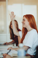 Lächelnde Studentin, die in der Klasse sitzt und die Hand hebt, um während der Vorlesung eine Frage zu stellen. Ein High-School-Student hebt die Hand und stellt dem Dozenten eine Frage. - JLPSF15202