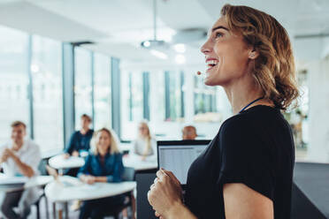 Fröhliche Geschäftsfrau bei einer Präsentation im Büro. Eine Unternehmerin steht am Podium und schaut lächelnd auf die Projektionsfläche. - JLPSF15203