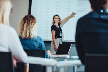 Geschäftsfrau, die eine Präsentation hält, während sie auf den Bildschirm zeigt, ihre Kollegen ansieht und lächelt. Weibliche Führungskraft, die während einer Konferenz eine informative Präsentation für ihr Team hält. - JLPSF15208