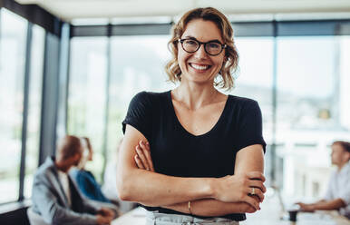 Portrait of a successful businesswoman standing with her arms crossed looking at camera and smiling. Positive female professional with colleagues sitting in meeting room at the back. - JLPSF15331