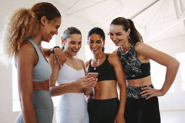 Nahaufnahme von Fitness-Frauen stehen zusammen nach dem Training und Blick auf ein Handy. Frauen, die Pause während des Trainings in einem Fitnessstudio Blick auf Smartphone und lächelnd. - JLPSF15361