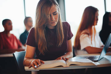 Studentin sitzt mit Laptop und Buch am Schreibtisch und lernt im Klassenzimmer einer High School. - JLPSF15374