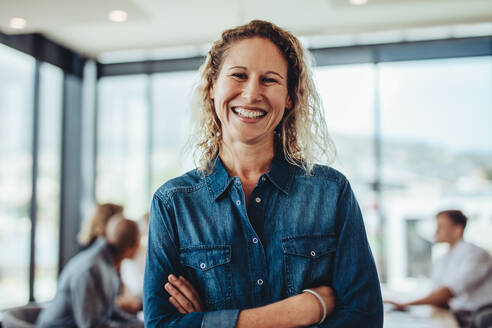 Portrait of smiling businesswoman standing in office with colleagues meeting in background. Successful female professional with her arms crossed. - JLPSF15409