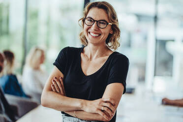 Portrait of smiling businesswoman standing in office with colleagues meeting in background. Successful female professional with her arms crossed in meeting room. - JLPSF15470