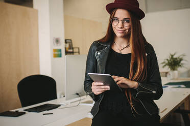 Lächelnde Geschäftsfrau mit Hut und Brille, die im Büro steht und einen Tablet-PC benutzt. Unternehmerin, die neben ihrem Tisch im Büro steht. - JLPSF15478