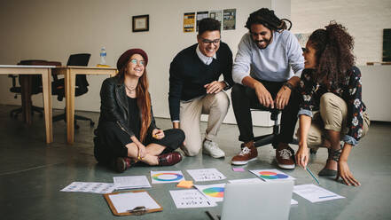 Geschäftspartner, die ihre Arbeit besprechen und dabei ihre Geschäftspapiere und Tabellen auf dem Boden arrangieren. Zufriedene Teamkollegen, die auf dem Boden sitzend mit Laptop und Geschäftsunterlagen ihre Arbeit besprechen. - JLPSF15488