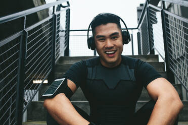 Porträt eines lachenden asiatischen Mannes, der auf einer Treppe sitzt und drahtlose Kopfhörer trägt, um Musik zu hören. Männlicher Sportler, der nach dem Training eine Pause macht. - JLPSF15500