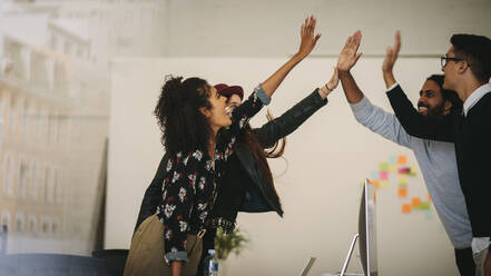 Geschäftskollegen, die sich im Büro stehend ein High Five geben. Teammitglieder, die ihren Erfolg bei der Arbeit feiern, indem sie ein High Five geben. - JLPSF15529