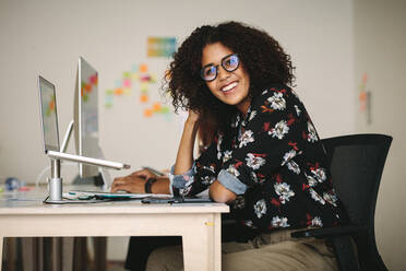 Smiling businesswoman sitting at her desk in office with a laptop mounted on a stand. Woman wearing eyeglasses working on laptop sitting in office. - JLPSF15535