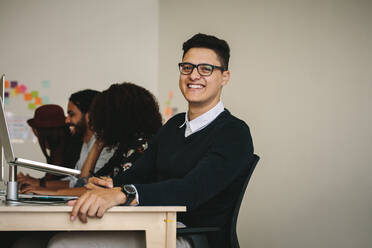 Ein lächelnder Geschäftsmann sitzt an seinem Schreibtisch mit einem Laptop auf einem Ständer. Ein Unternehmer sitzt zusammen mit seinen Kollegen im Büro. - JLPSF15539