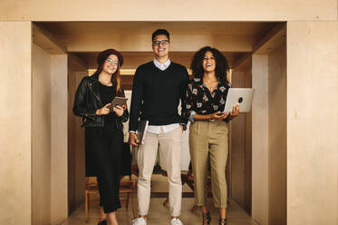 Happy teammates standing in office holding laptops and a tablet. Businessman with two female colleagues at work standing holding laptops. - JLPSF15543