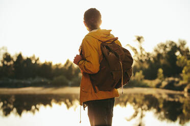 Mann mit Rucksack steht an einem See und schaut in die Abendsonne. Tourist erkundet die Natur im Freien an einem See mit Sonne im Hintergrund. - JLPSF15566