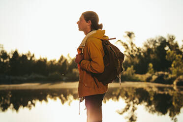 Side view of a man wearing backpack standing beside a lake and looking away. Traveler standing near a lake and looking around with sun in the background. - JLPSF15567
