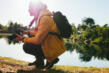Seitenansicht eines Reisenden, der an einem See sitzt und eine Digitalkamera in der Hand hält. Ein Mann, der an einem sonnigen Tag an einem See sitzt und Fotos von der Natur macht. - JLPSF15573