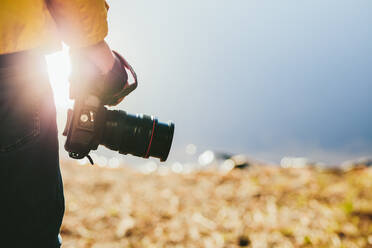 Ausschnitt eines Mannes, der eine DSLR-Kamera in der Hand hält. Nahaufnahme der Hand einer Person, die eine Digitalkamera hält, mit Sonnenflecken im Hintergrund. - JLPSF15576