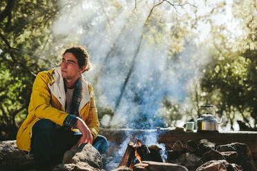 Junger Mann in einer ländlichen Umgebung, der an einem Lagerfeuer sitzt, mit einem Kessel und einer Tasse im Hintergrund. - JLPSF15581