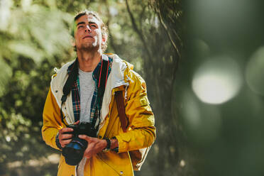 Porträt eines Mannes mit Jacke und Rucksack, der in einem Wald spazieren geht und eine DSLR-Kamera hält. Ein Mann, der einen Wald erkundet und die Schönheit mit einer Digitalkamera festhält. - JLPSF15582