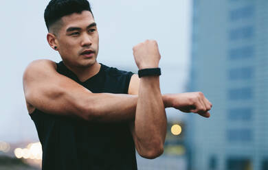 Fit young man with muscular build standing outside stretching hands and looking away. Asian fitness model doing warmup workout. - JLPSF15588