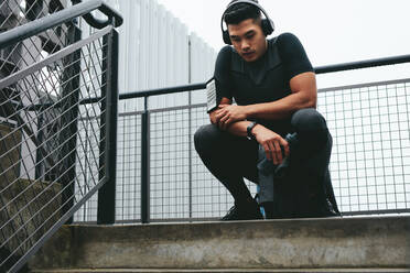 Fitness man relaxing after workout sitting outdoors. Male athlete listening to music using wireless headphones and relaxing after outdoor training session. - JLPSF15591